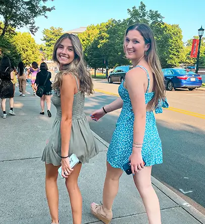 two young women turn to pose for a photo while walking on the sidewalk
